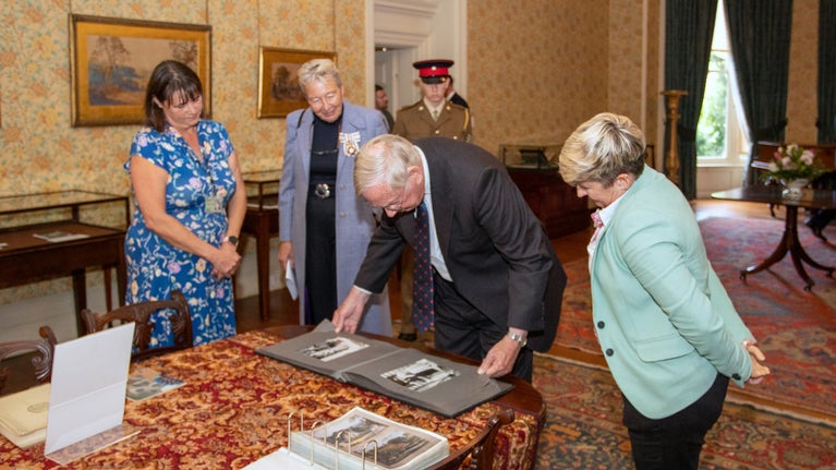 His Royal Highness The Duke of Gloucester is looking at a photo album on a table, surrounded by three ladies and a man in military uniform